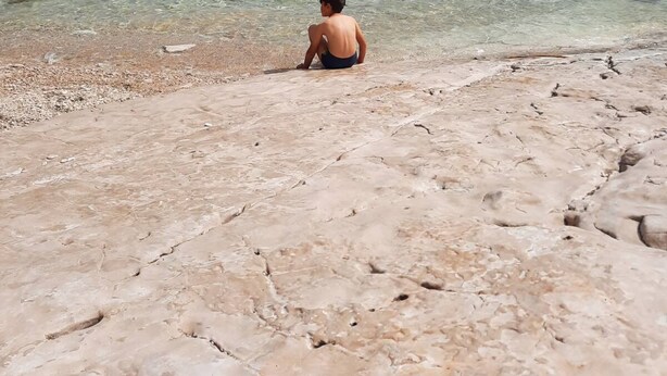 Una persona seduta su una spiaggia rocciosa con acqua limpida e una costa in lontananza.