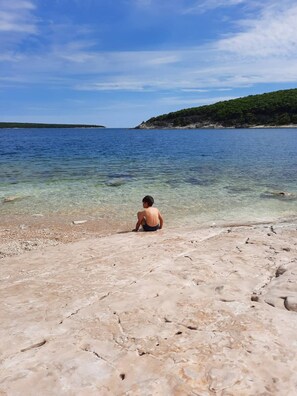 Eine Person sitzt auf einem felsigen Strand mit klarem Wasser und einer entfernten Küstenlinie.