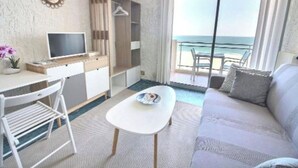 A room with a sofa, a small table, a TV, a desk, and a chair. There is a balcony with a view of the sea. - Studio au Pied de la Plage (Canet-en-Roussillon)