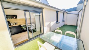 Een klein terras met een glazen tafel en stoelen, een schuifpui naar een keuken en kunstgras.