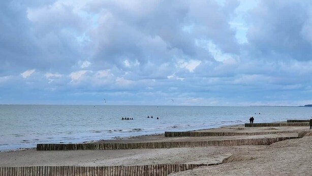 Een strand met een houten hek, rustig water en een bewolkte lucht.