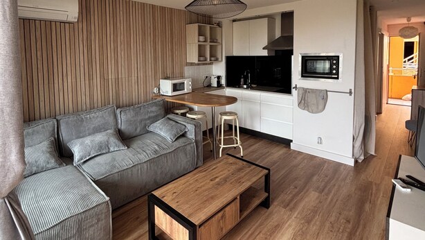 Een moderne woonkamer met een hoekbank, een houten salontafel en een keukenhoek met witte kasten en een magnetron.