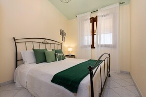 Un lit bien fait, agrémenté d’un plaid vert, une lampe de chevet et une fenêtre ornée de rideaux légers.