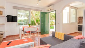 Una sala moderna con un sofá gris, una mesa de comedor con sillas, un televisor de pantalla plana montado en la pared y una cocina visible a través de una puerta abierta.