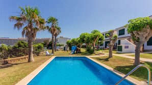 Una piscina con una escalera metálica, rodeada de palmeras y un tobogán, con edificios al fondo.