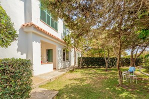Un bâtiment blanc avec un toit en tuiles, un porche abrité et un petit jardin agrémenté d’un banc et de chaises.