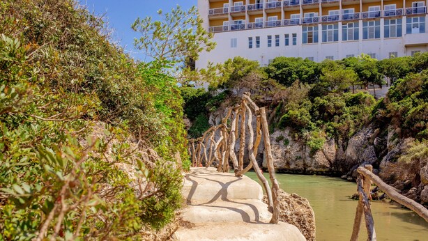 Una escalera de piedra conduce a un edificio con balcones, rodeado de vegetación y una masa de agua.