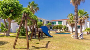 Un parque infantil con tobogán y columpios, rodeado de palmeras y un edificio con techo de tejas rojas.