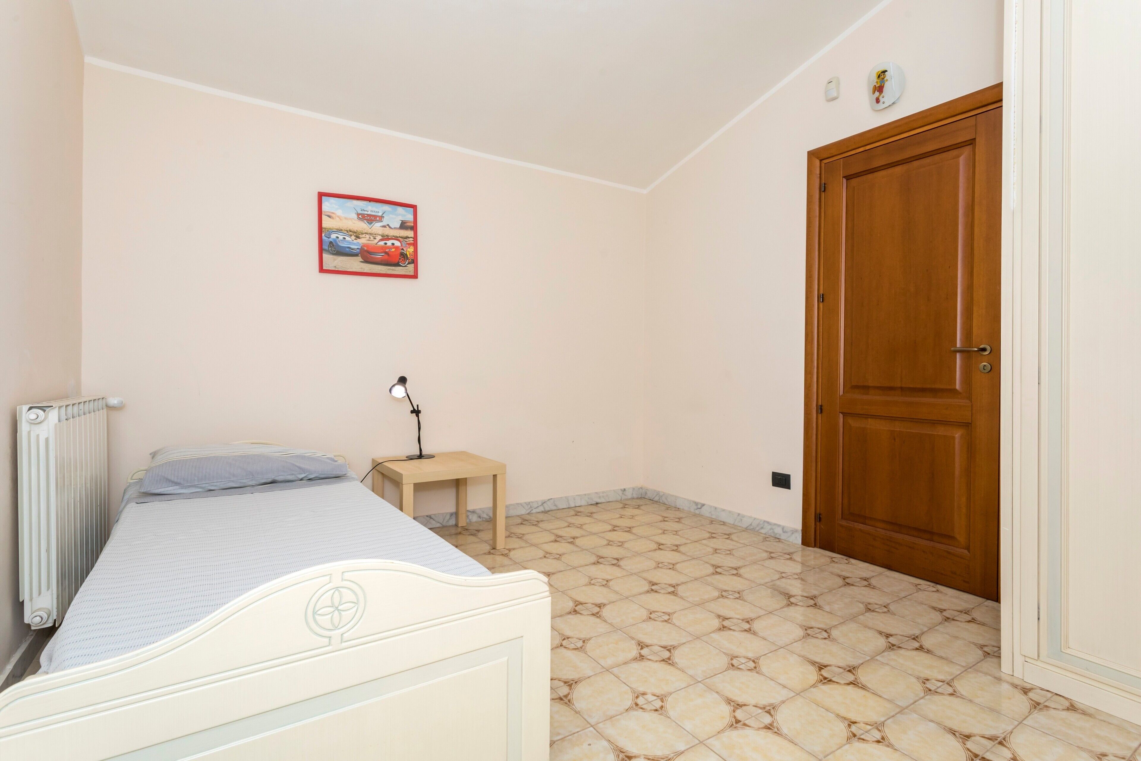 Un dormitorio con cama, mesita de noche, lámpara, radiador, puerta y un cuadro colgado en la pared.