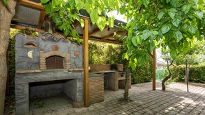 Une structure extérieure en briques abritant un four à bois, surmontée d’une pergola en bois et entourée de verdure.