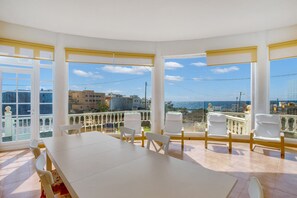 Um quarto com uma mesa grande e cadeiras, janelas amplas com persianas amarelas e vista para o mar e edifícios.