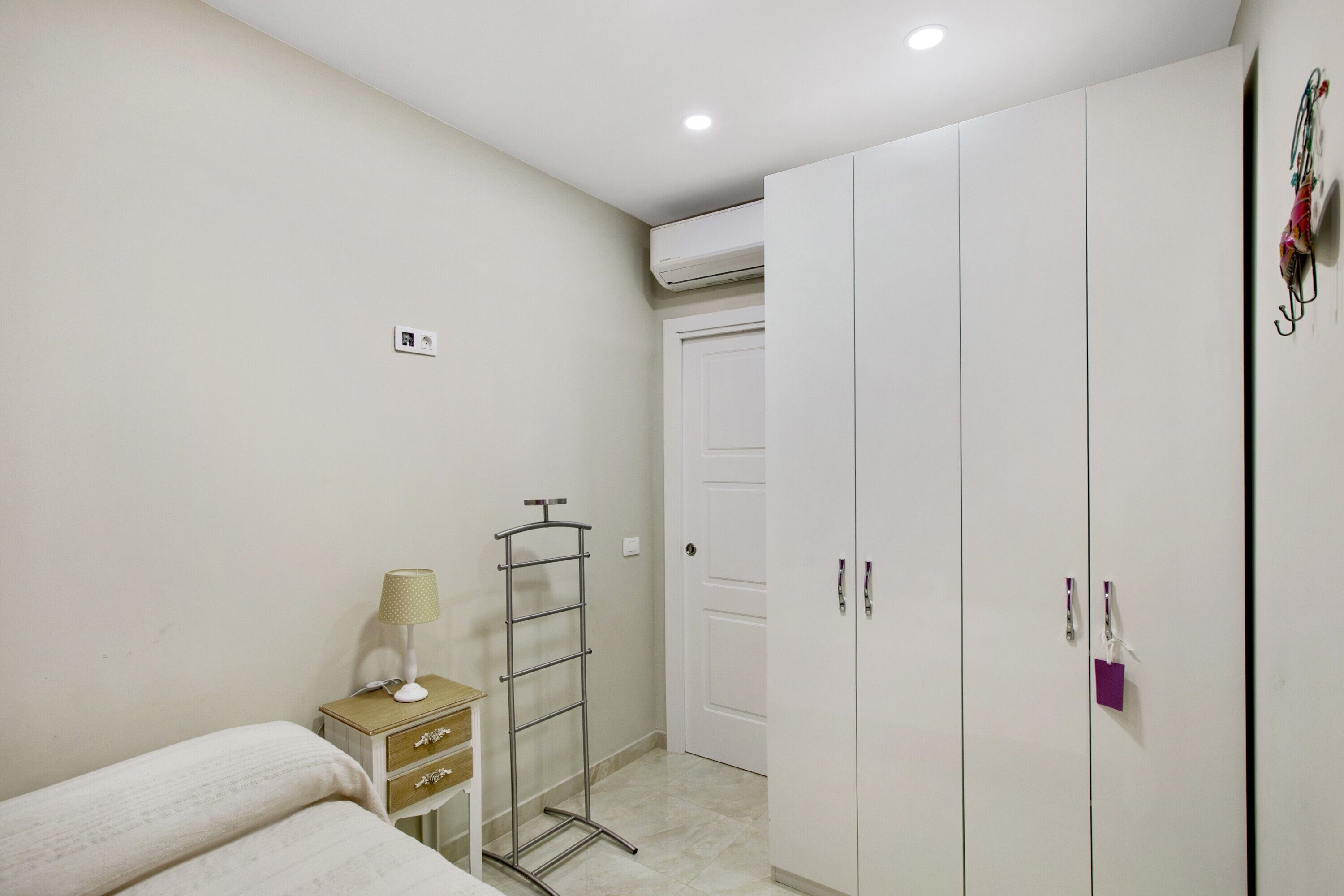 A bedroom with a bed, a bedside table with a lamp, a wardrobe, and a hanging clothes rack.