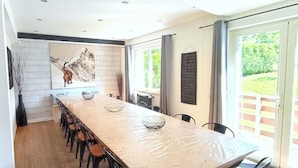 A modern kitchen with a long dining table, chairs, a decorative wall piece, and a view of greenery through large windows.