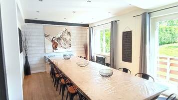 Une cuisine moderne avec une longue table à manger, des chaises, un élément décoratif mural et une vue sur la verdure grâce à de grandes fenêtres.
