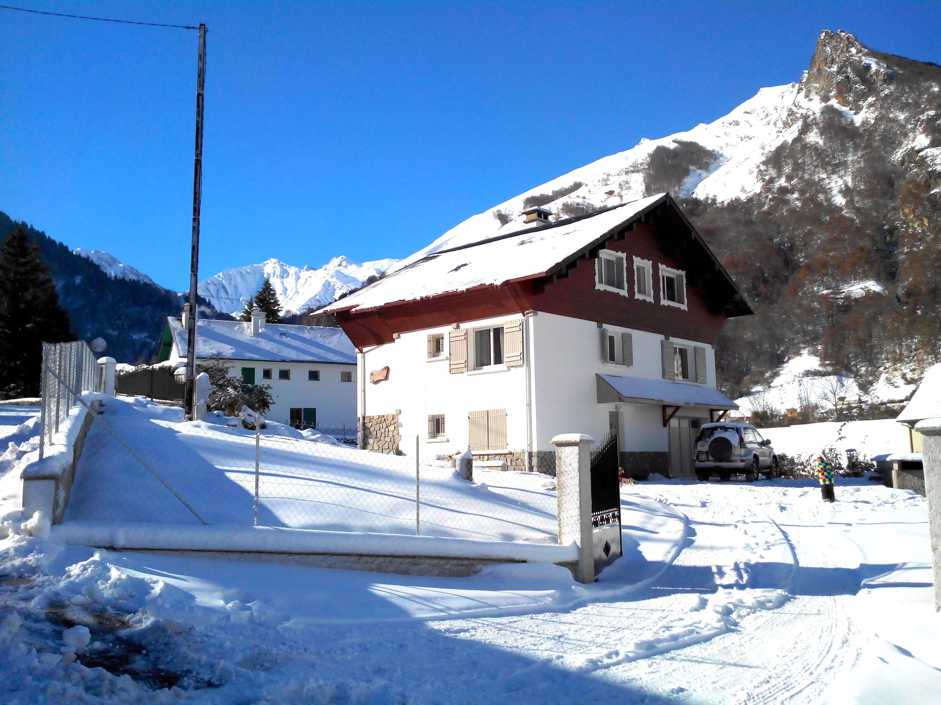 Un pueblo cubierto de nieve en la montaña, con una casa destacada que tiene techo rojo y paredes blancas, camino empedrado y cielo azul despejado.