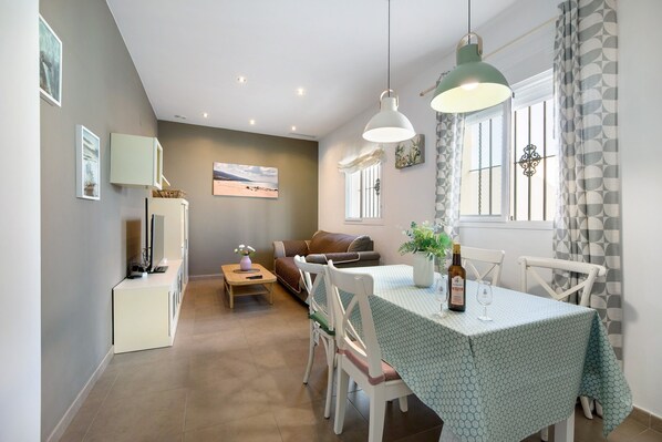 Un espace repas comprenant une table, des chaises, un luminaire suspendu, une fenêtre avec des rideaux et un canapé à l’arrière-plan.