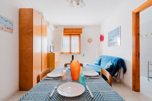 A dining area with a table set for two, a wooden wardrobe, a window with curtains, and a bed with a blue cover.