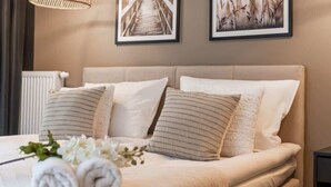 A neatly made bed with rolled towels and white flowers, a radiator, and two framed pictures on the wall.