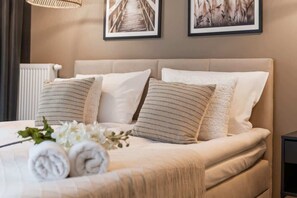 A neatly made bed with rolled towels and white flowers, a radiator, and two framed pictures on the wall.