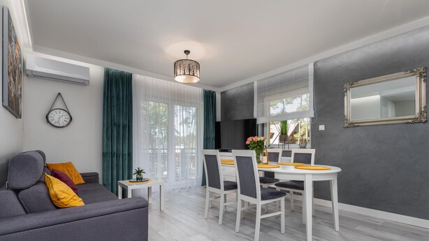 Un salon moderne avec un canapé gris, une table à manger avec des chaises blanches et un coin cuisine agrémenté d’un grand miroir et de murs gris.