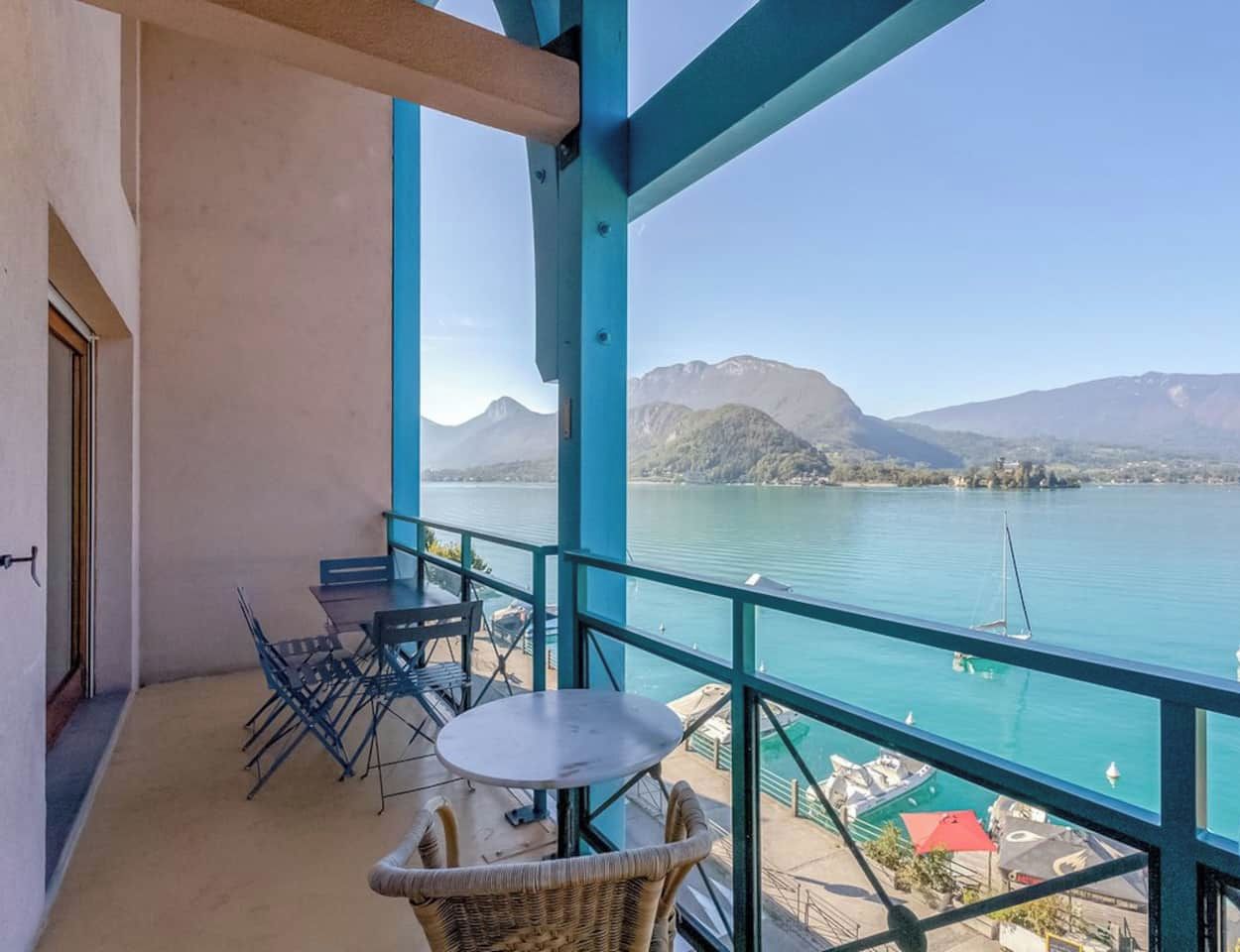 A balcony with a table and chairs, overlooking a lake with boats and mountains in the background.