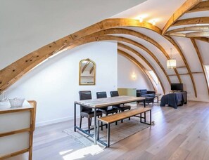 Et rummeligt loftsværelse med højloftet loft og synlige træbjælker. Værelset har et spisebord med stole, en sofa, et fjernsyn og et stort spejl på væggen.