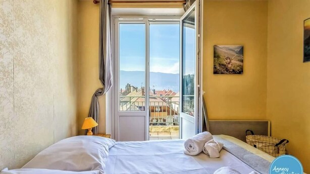 Ein Hotelzimmer mit Bett, einem fenster mit Vorhängen, einer Lampe, einem Gemälde an der Wand und Blick auf Gebäude.