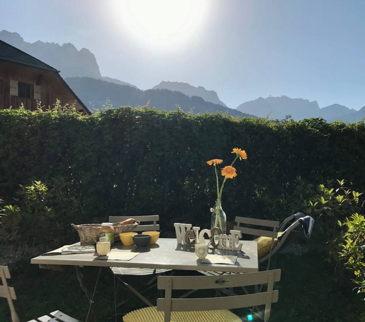 Uma área de refeições ao ar livre com uma mesa posta para dois, cercada por um viveiro e um cenário de montanhas.