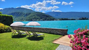 Un espace aménagé au bord de la piscine, avec des chaises longues et des parasols, un mur en pierre et une vue sur un lac, avec des montagnes en arrière-plan.