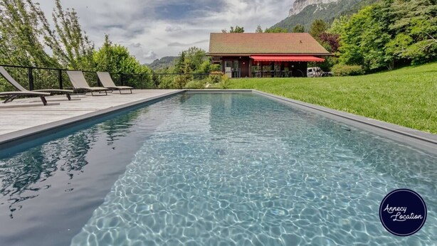 Una piscina rettangolare con acqua cristallina, circondata da un solarium in legno con lettini. Sullo sfondo, un edificio con tetto rosso e una catena montuosa.