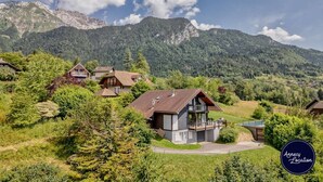 Un villaggio di montagna con case in legno tradizionali, un'abitazione moderna centrale con un balcone, e vista chiara sulle montagne circostanti.