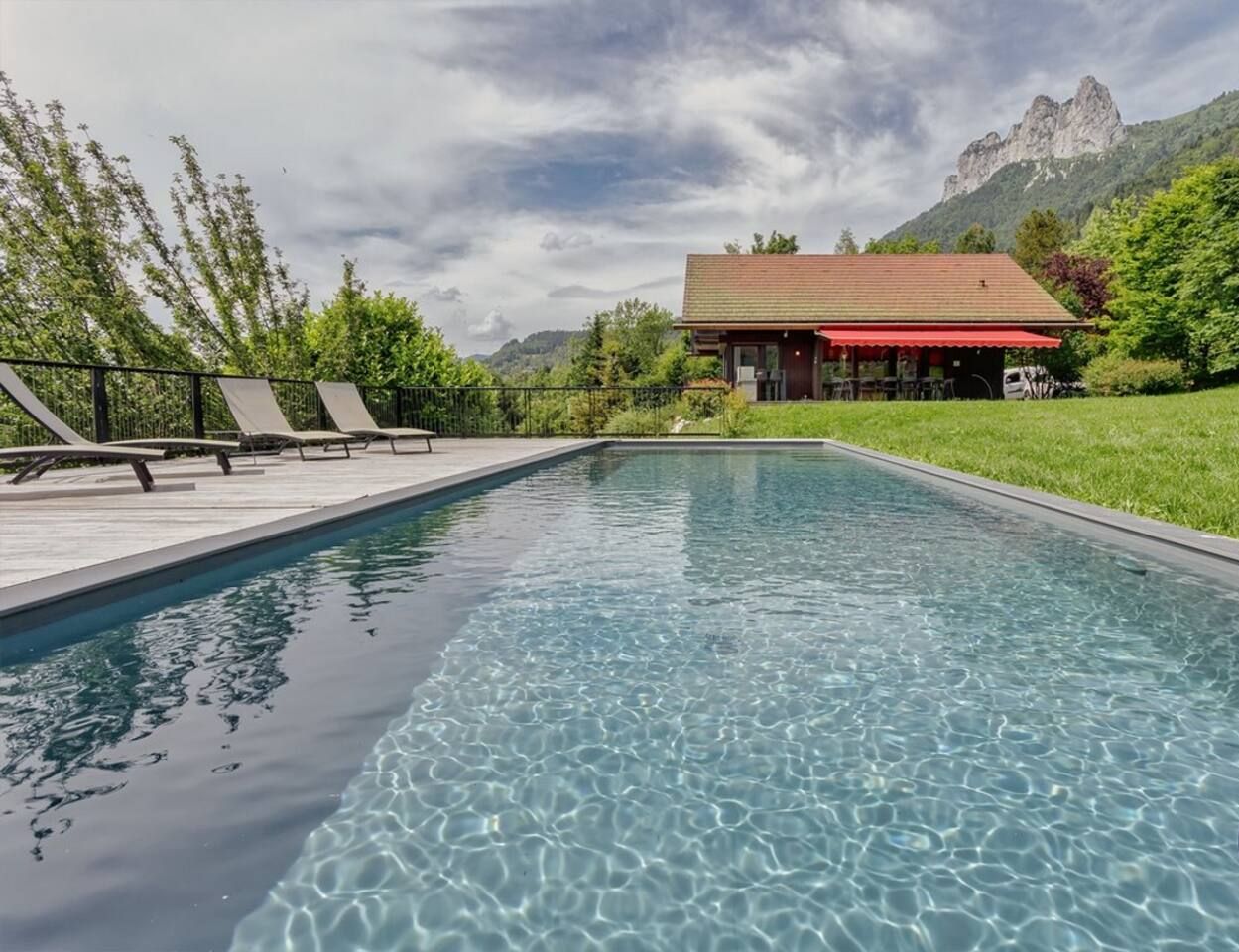 A rectangular swimming pool with clear water, surrounded by a wooden deck with lounge chairs. In the background, there is a building with a red roof and a mountain range.