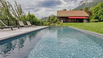 A rectangular swimming pool with clear water, surrounded by a wooden deck with lounge chairs. In the background, there is a building with a red roof and a mountain range.
