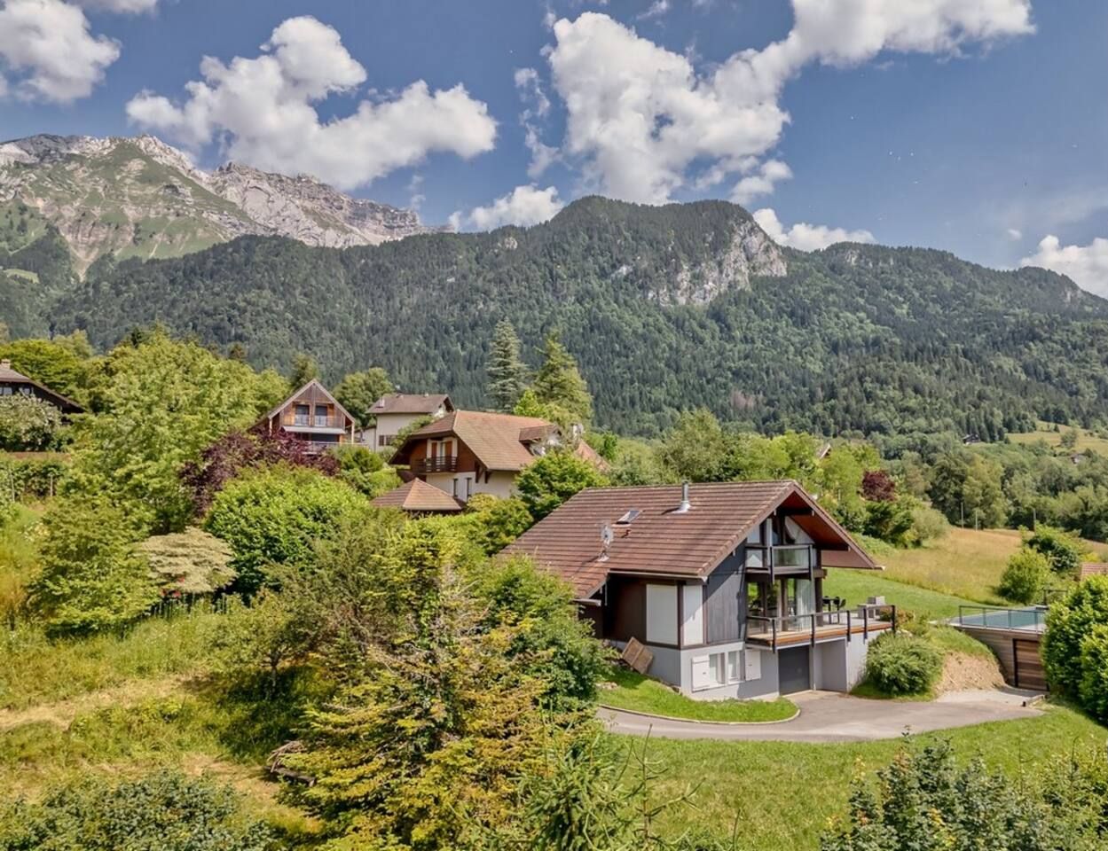 A mountain village with traditional wooden houses, a central modern house with a balcony, and a clear view of the surrounding mountains.