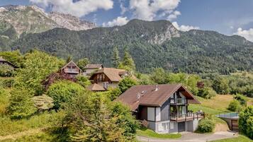 A mountain village with traditional wooden houses, a central modern house with a balcony, and a clear view of the surrounding mountains.