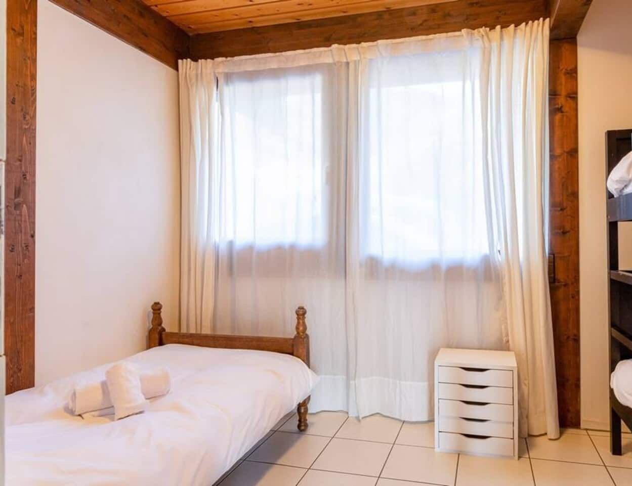 Ein Schlafzimmer mit einem hölzernen Bett, einem kleinen Nachttisch, einem Etagenbett und einem Fenster mit transparenten Vorhängen.