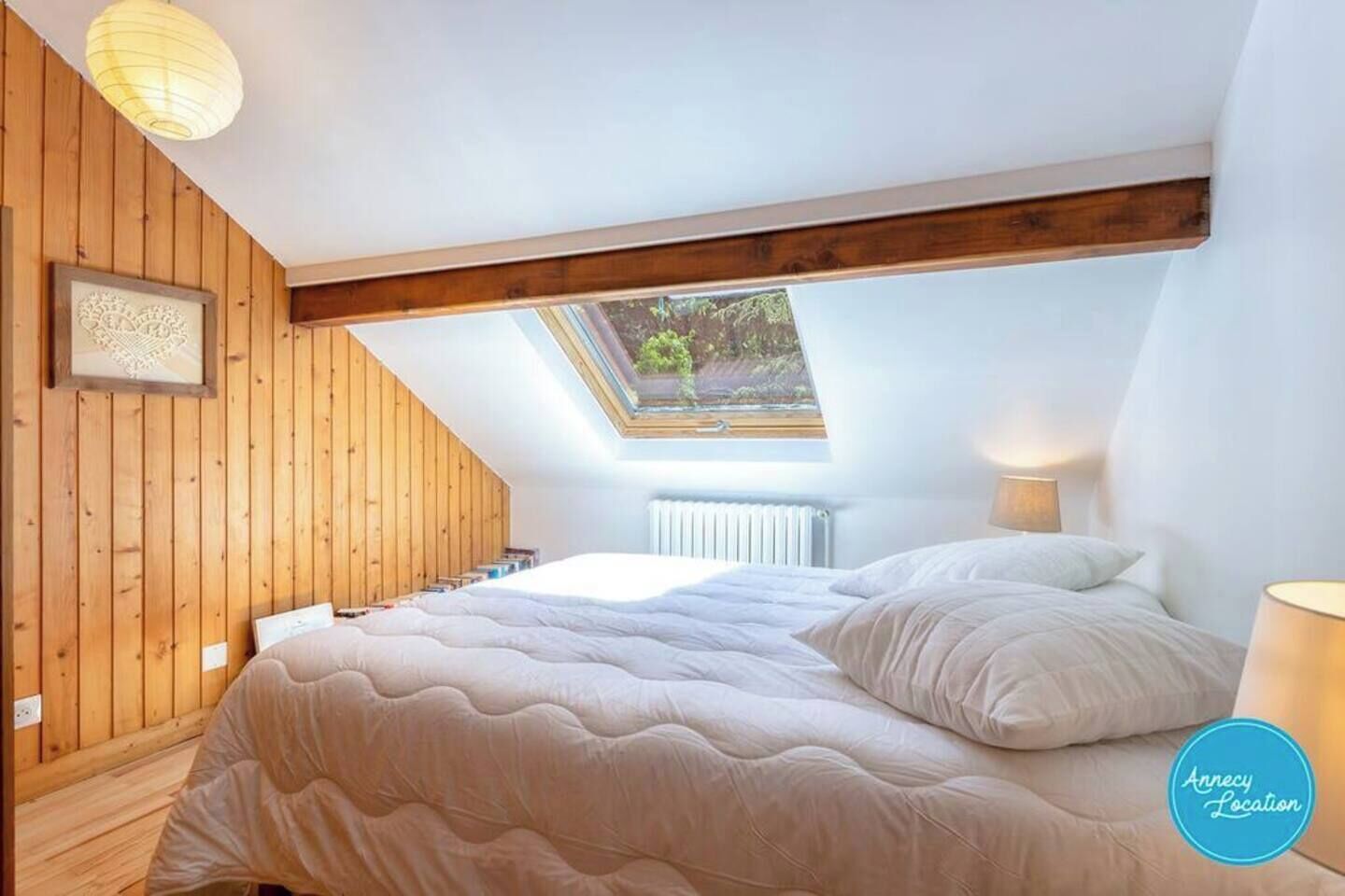 Ein Schlafzimmer mit schrägen Wänden, Holzwänden, einem Oberlicht, einem Bett mit weißer Bettwäsche und einer Nachttischlampe.