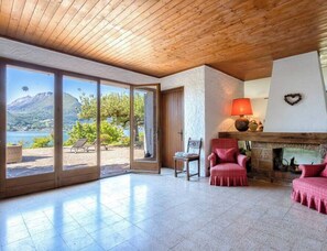 Il s’agit d’une pièce spacieuse dotée d’une grande porte-fenêtre coulissante qui donne sur un balcon offrant une vue sur les montagnes. À l’intérieur, on trouve une cheminée avec une lampe rouge, deux fauteuils rouges et une petite table avec une chaise.