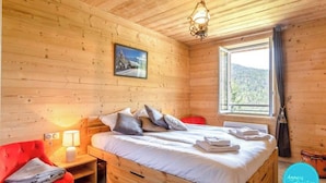 Ein Schlafzimmer einer Holzhütte mit einem Bett, einem roten Sessel, einem Nachttisch mit einer Lampe und einem Fenster mit Blick ins Freie.