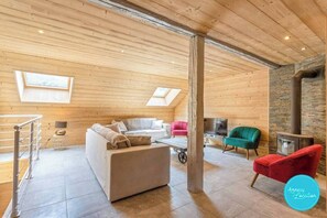 Una habitación tipo loft acogedora con techo de madera, pared de piedra y chimenea. Hay un sofá, una silla roja y un televisor.