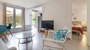 A modern living room with a flat-screen TV, a round table, and a wicker chair with a leaf-patterned pillow. - Suite Familiale - Le Clos Vallis (Sarlat-la-Canéda)