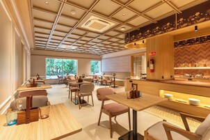 A modern restaurant with a high ceiling, wooden tables, and chairs. There is a fire extinguisher on the wall and a window providing a view of trees.