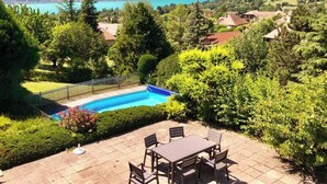 A pool with a view of a lake and mountains, surrounded by greenery and a patio with chairs.