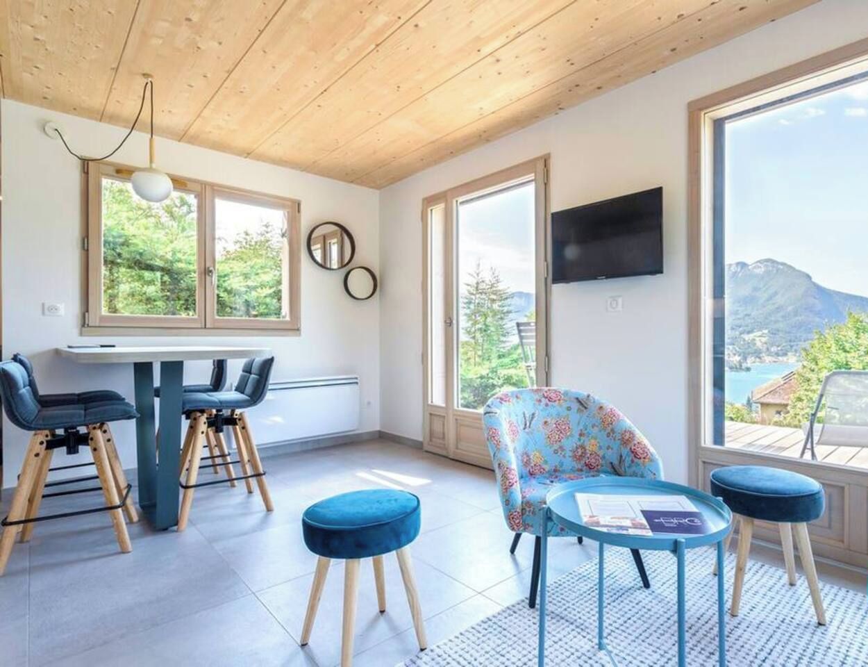 Een moderne kamer met een houten plafond, grote ramen, een televisie, een tafel met stoelen en uitzicht op een bergachtig landschap.