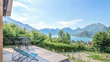 Una terraza de madera con muebles al aire libre, con vista a un paisaje montañoso y un lago.