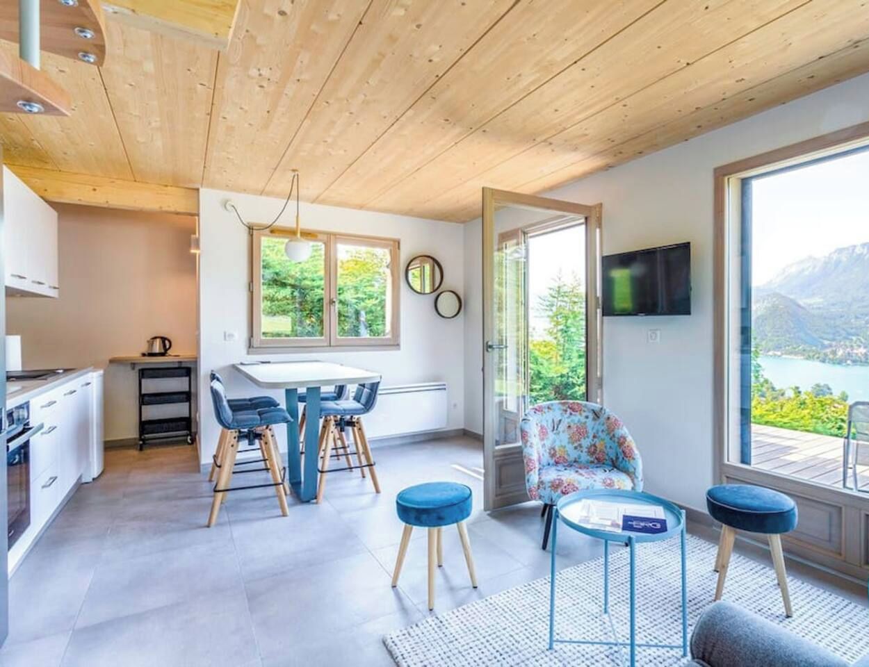 Una cocina moderna con armarios blancos y un comedor con una mesa y sillas. Hay un televisor en la pared y vistas a montañas y un lago a través de ventanas amplias.