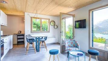 Una cocina moderna con armarios blancos y un comedor con una mesa y sillas. Hay un televisor en la pared y vistas a montañas y un lago a través de ventanas amplias.