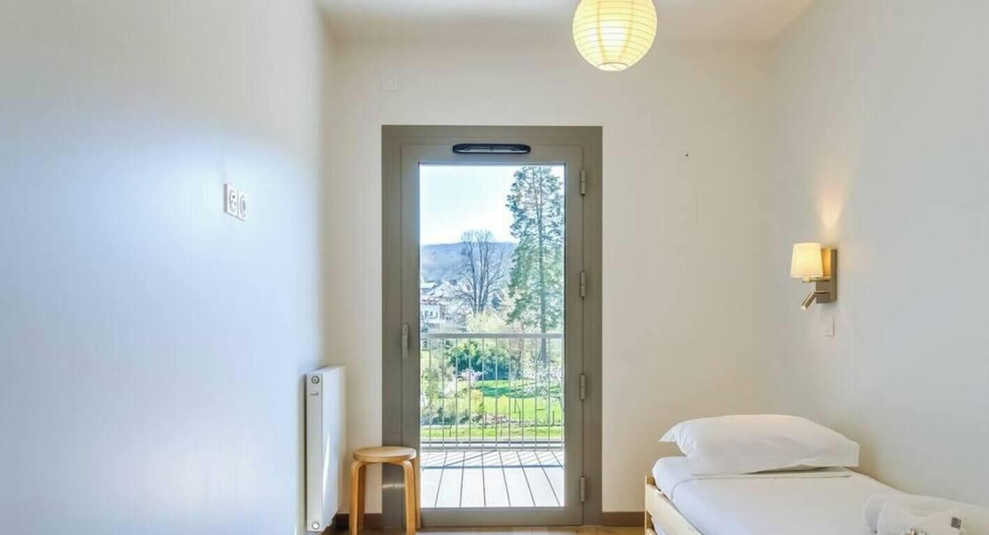 A bedroom with a bed, a small stool, a radiator, a balcony door, and a view of the outdoors.