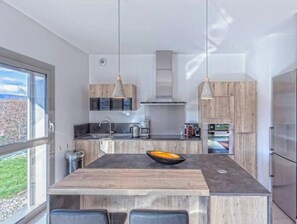 A modern kitchen with a central island, pendant lighting, and a view through large windows.