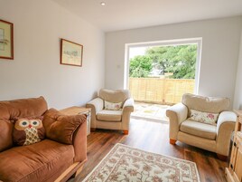 A living room with a brown leather sofa, two beige armchairs, a wooden coffee table, and a patterned rug. There are two framed pictures on the wall and a large window with a view of greenery outside.